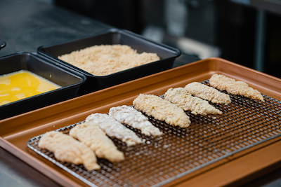 Raw, breaded chicken tenders on a drying rack, with a plate of egg and a plate of bread crumbs next to it.