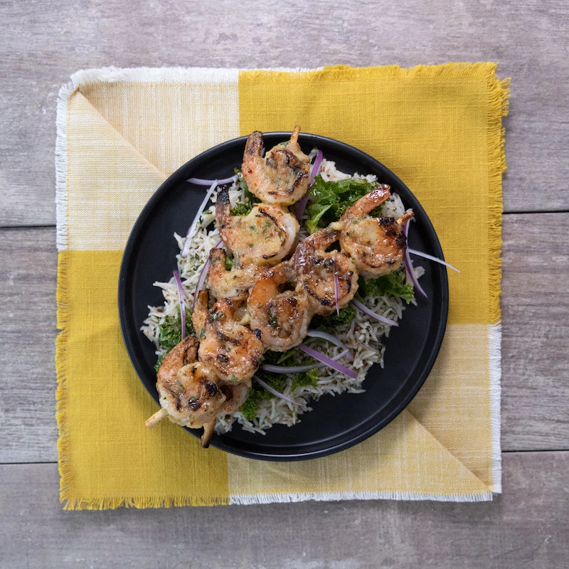 Grilled shrimp on skewers, glazed with a mango and habanero sauce. They sit on a bed of rice on a black plate, with a yellow napkin underneath. 