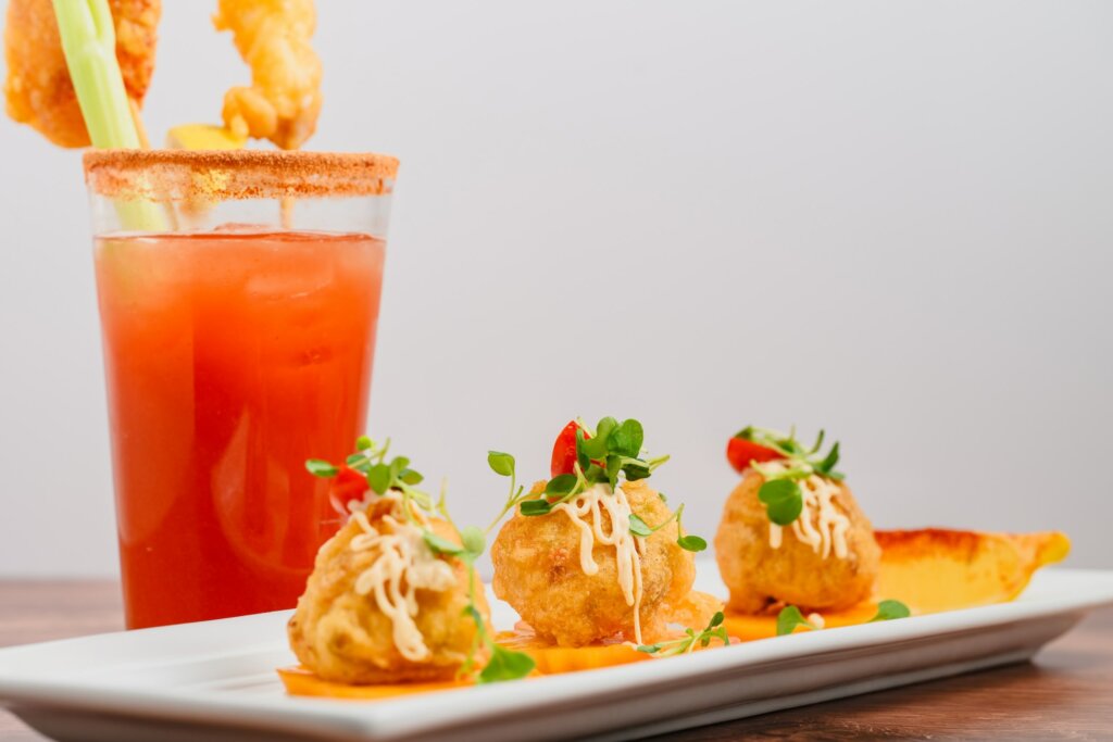 A restaurant-branded appetizer featuring crispy, custom-flavored bites topped with microgreens and sauce, served alongside a visually branded Bloody Mary cocktail.