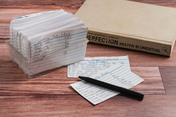 Recipe cards neatly organized in a plastic holder, accompanied by a pen and a culinary book.