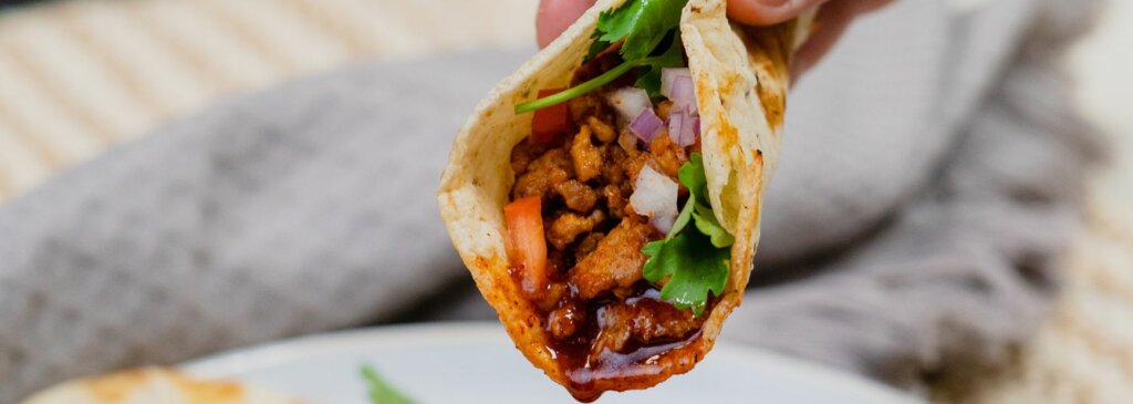 A hand holding a soft corn tortilla taco filled with seasoned ground meat, chopped tomatoes, diced red onions, and fresh cilantro. The taco is positioned above a small bowl of rich, reddish-brown dipping sauce, birria consommé. In the background, another taco rests on a white plate atop a textured beige and gray surface, slightly out of focus.