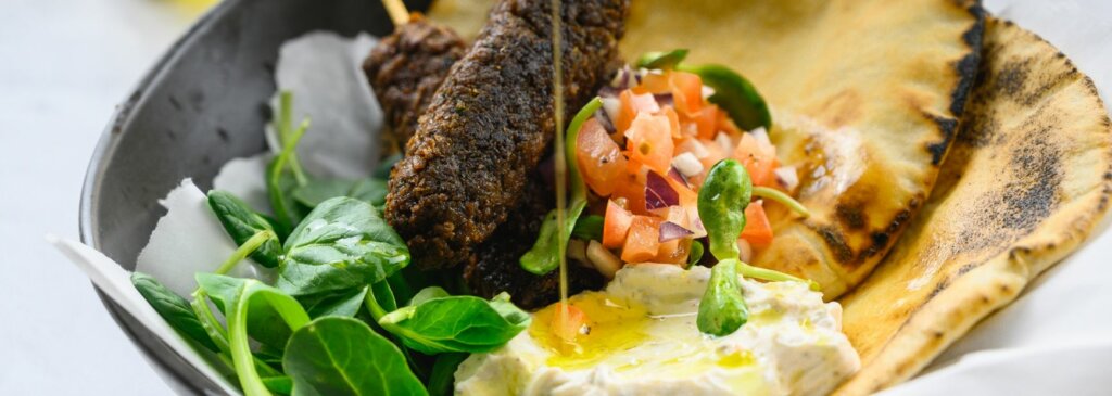 Bowl featuring grilled pita, falafel skewers, creamy hummus drizzled with olive oil, fresh spinach leaves, and a topping of diced tomatoes and red onions, arranged in a modern presentation.