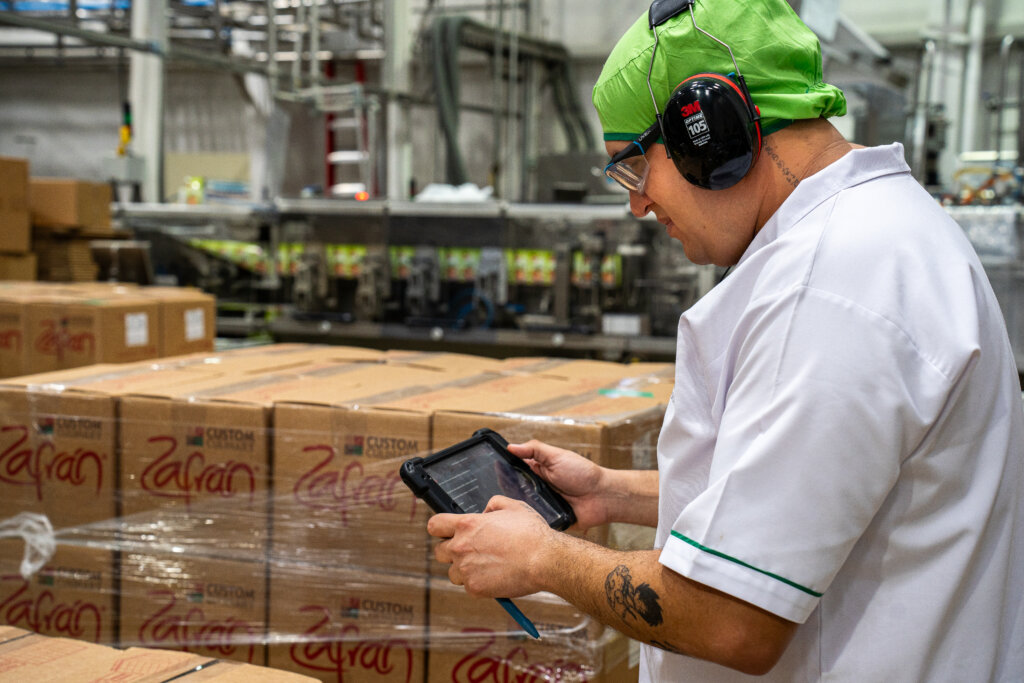 Factory worker using a tablet to inspect and verify food supply shipments in a warehouse filled with ingredient boxes.